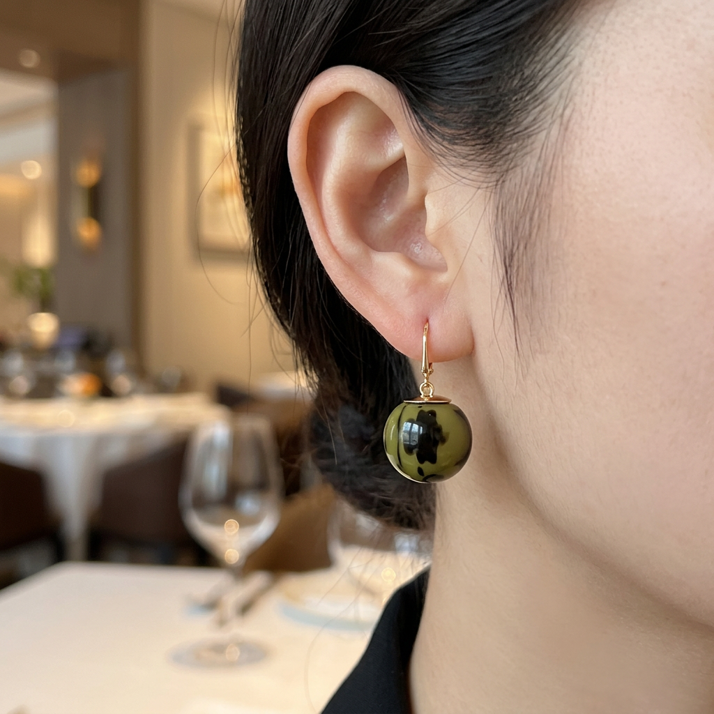 Kolczyk ze złotym zapięciem i zielono-czarną kulką na modelce w restauracji